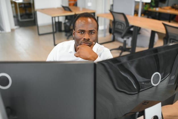 A person of color looking thoughtfully at their computer screen in an office setting. The lighting is soft and professional, with a focus on the individual's expression, conveying resilience and determination. The background includes blurred office elements like bookshelves and other colleagues.