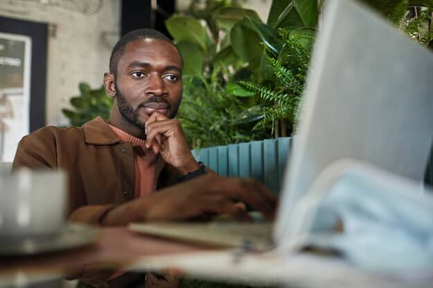 A person of color sitting at their desk, looking thoughtfully at their computer screen with a concerned expression. Surrounding them are blurry images of coworkers, who all seem to be in a conversation with each other or absorbed in their own work.