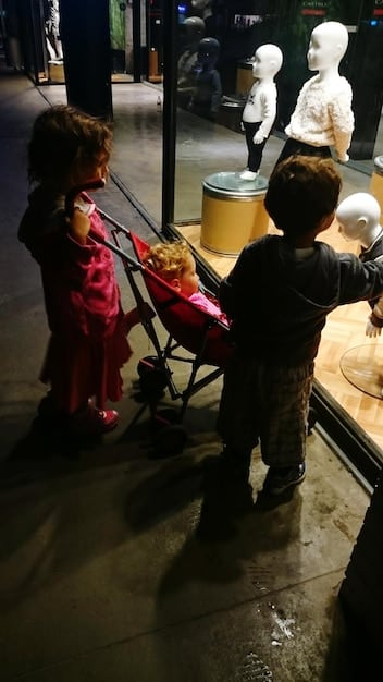 A wide shot of an immersive theater performance in a warehouse, showing actors interacting closely with audience members who are dressed in period costumes. The lighting is moody and atmospheric.