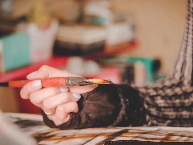 A close-up of an artist's hands working on an abstract painting in their studio. The artist is using vibrant colors and expressive brushstrokes. There are various tools like brushes, palettes, and jars around.